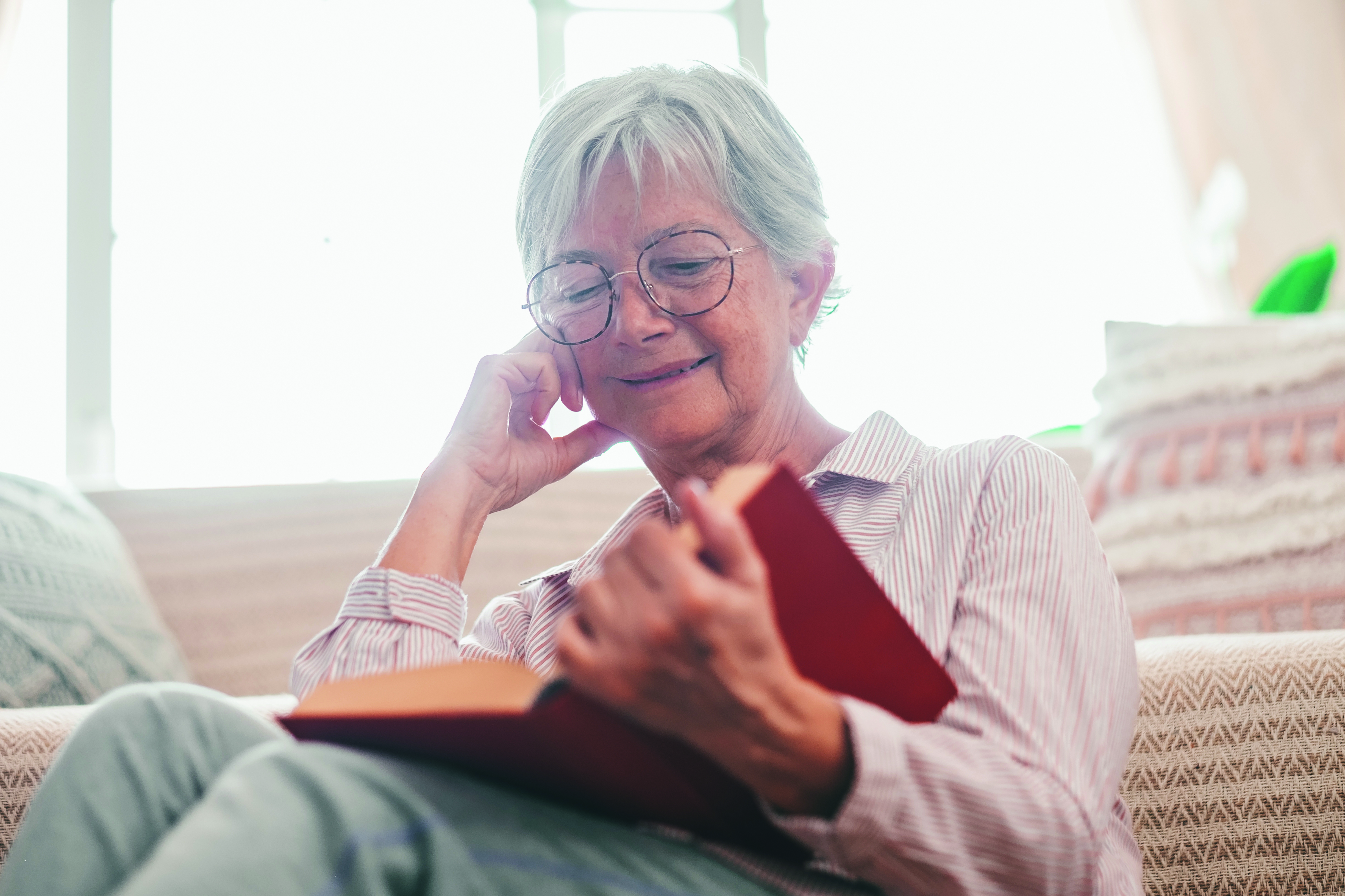 An elderly smiling woman, with glasses and short gray hair, is sitting comfortably on a sofa while reading a book in a cozy, naturally lit setting.